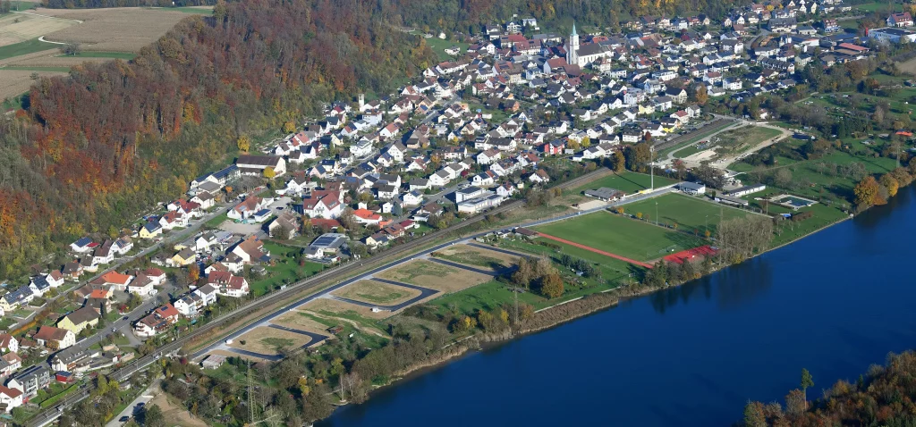 Eine Luftaufnahme eines Dorfes oder einer Kleinstadt, umgeben von Natur, mit einem Fluss im Vordergrund und einem Baugebiet in der Nähe.